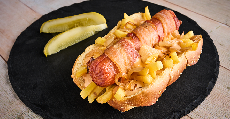A bacon-wrapped hot dog topped with fries and grilled onions, served with pickles on a black plate