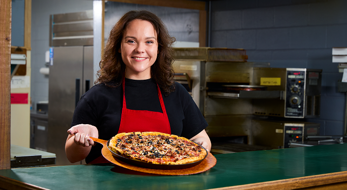 A smiling woman in a red apron holds a deep-dish pizza on a wooden peel in a restaurant kitchen