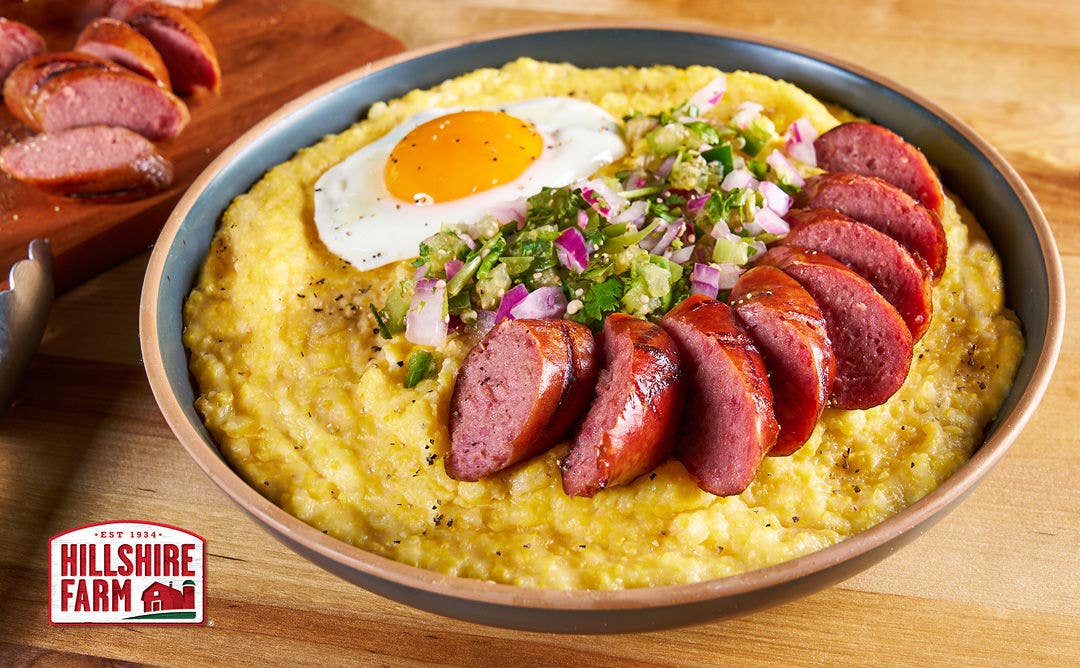 A bowl of cheddar grits topped with a sunny-side-up egg, sliced smoked sausage and pico de gallo  