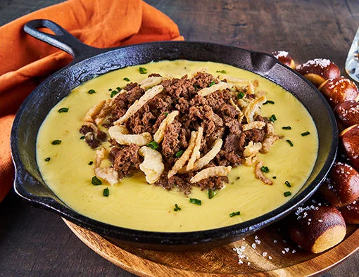 Warm smoked gouda fondue skillet with tender sliced beef steak topped with crispy fried shallots and chives served with salted pretzel bites