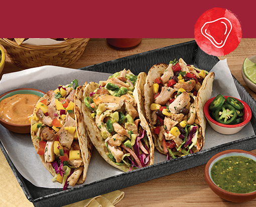 Tray of three smoked chicken tacos topped with cabbage slaw and vegetables, served with jalapeños, salsa verde, and creamy sauce on wooden table
