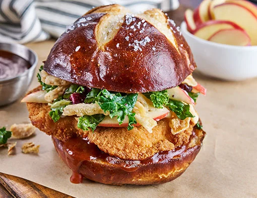 Smoky burnt honey BBQ chicken sandwich with crispy Tyson breaded chicken filet, apple kale slaw, and fried onion strings on a salted pretzel bun