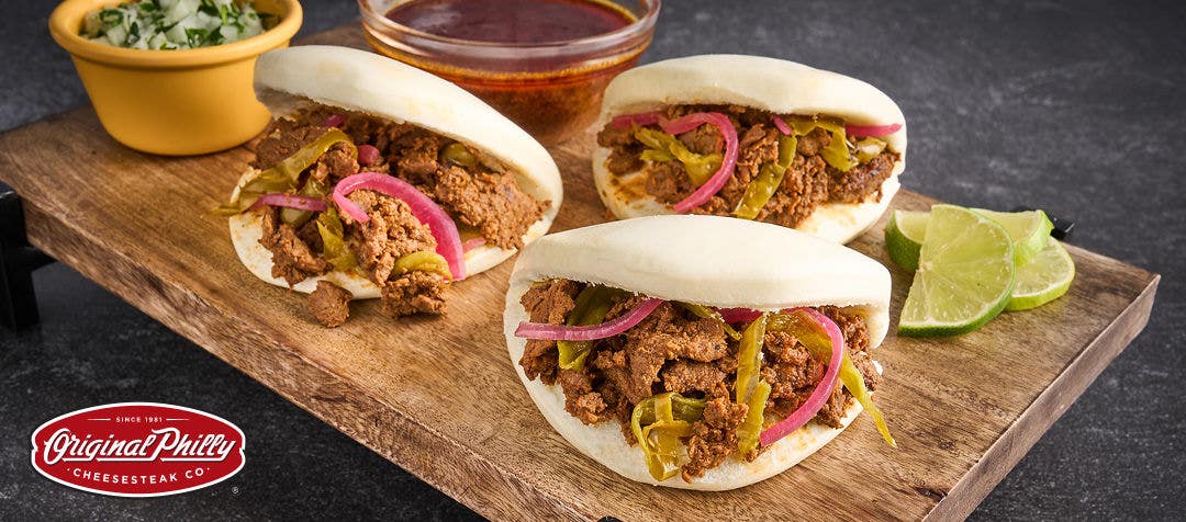 Three small slider-style sandwiches filled with sliced beef, pickled onions, cilantro and served on a cutting board with sliced lime and sauces on the side  