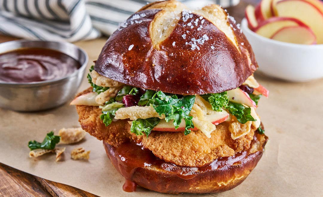 A fried chicken sandwich on a pretzel bun loaded with fried onions and slaw with side dishes in the background 