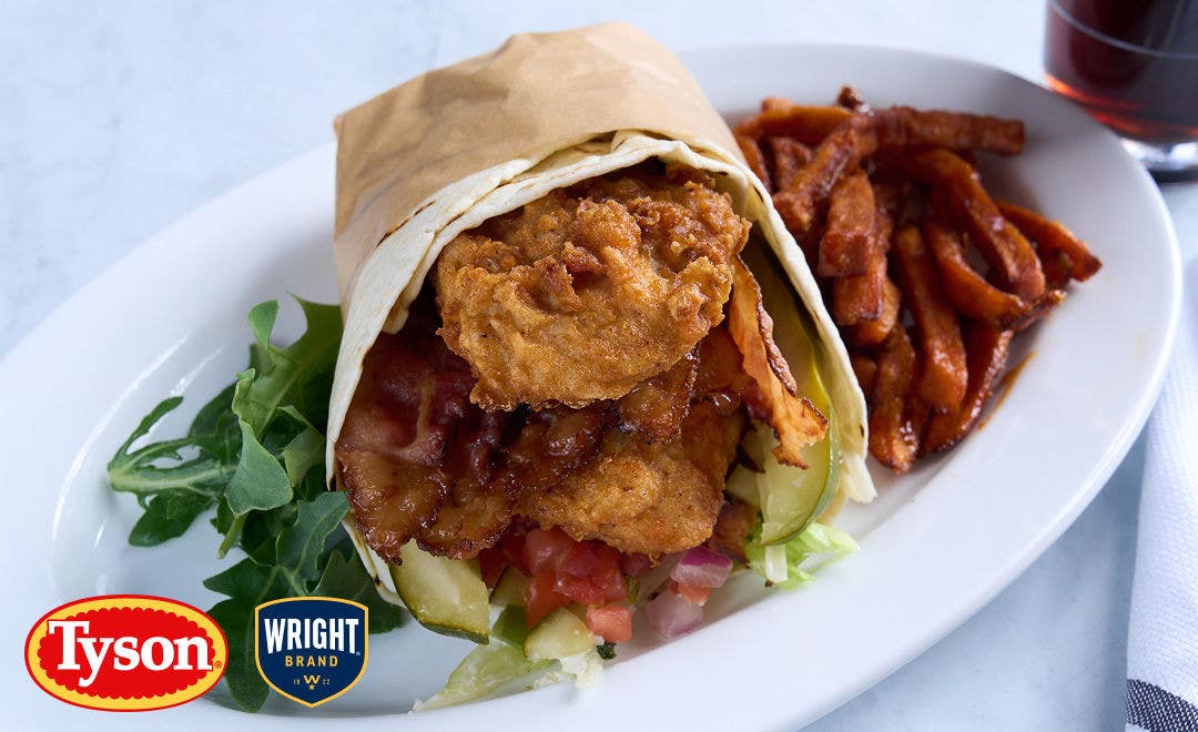 A white plate showing the open end of a flour tortilla wrapped in parchment and stuffed with breaded chicken tenderloins, cheese, pickle spears and lettuce with a side of fries 