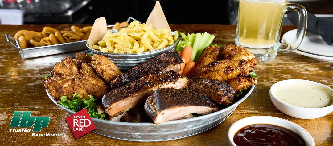 A table setting with a large platter of wings and barbecue ribs, two side sauces and onion rings with a mug of beer in the background   