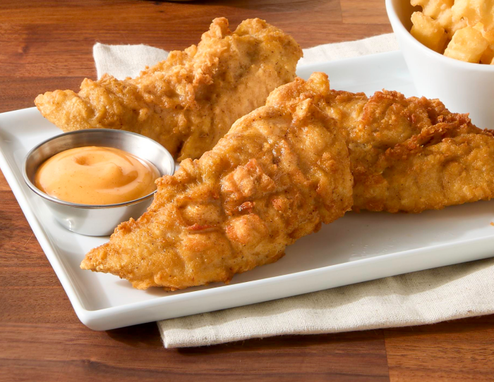 Two honey-flavored breaded chicken tenderloins on a white plate with a cup of honey dipping sauce, served with fries on a wooden table