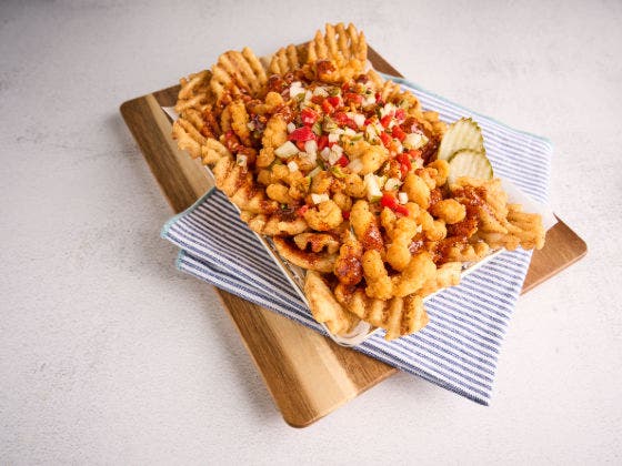 Waffle fries topped with crispy chicken bites, red sauce, relish, and pickles in a basket