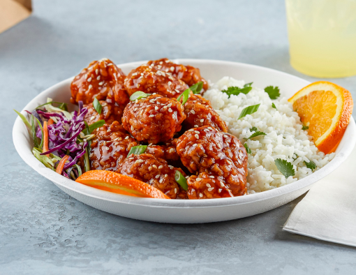 A bowl of breaded chicken chunks in sauce served over slaw and white rice garnished with orange slices 
