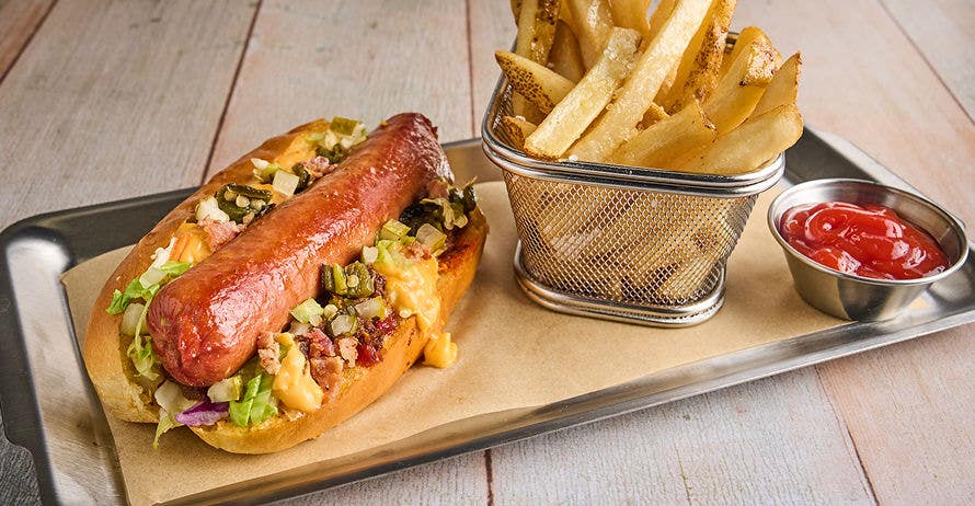 A loaded hot dog with cheese, onions, and relish on a metal tray with fries and ketchup