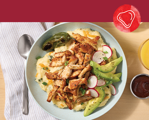 Bowl of creamy grits topped with smoked chicken pieces, sliced avocado, radishes, roasted pepper, and green onions on a light wooden table