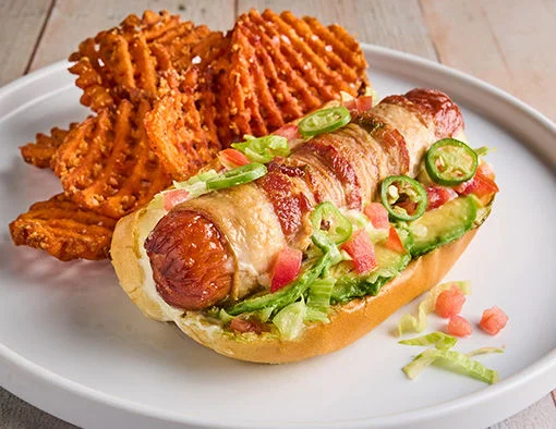 A bacon-wrapped hot dog topped with avocado, lettuce, tomatoes, and jalapeños, served with sweet potato fries