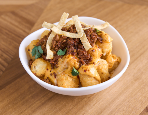 Potato tots and beef taco meat in a white bowl covered in nacho cheese, creamy sauce, fresh cilantro and tortilla strips 