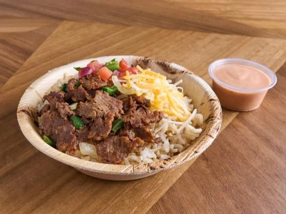 Bowl with seasoned steak, rice, poblano peppers, shredded cheese, fresh veggies, and a side of sauce