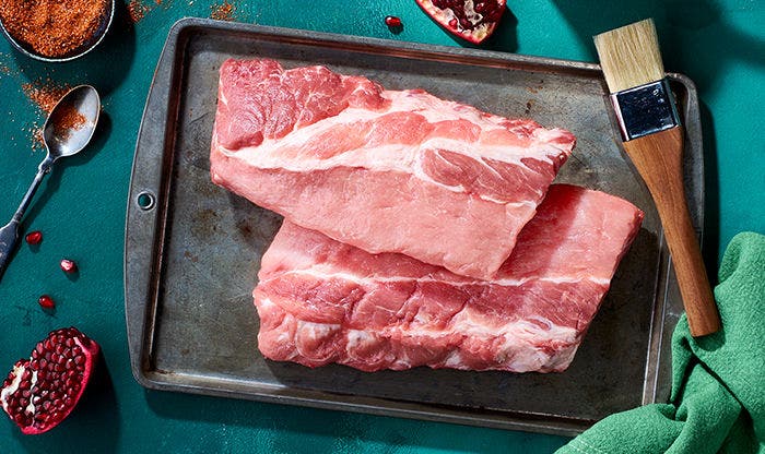Raw pork ribs on a tray, halved pomegranate, seasoning spoon, and basting brush