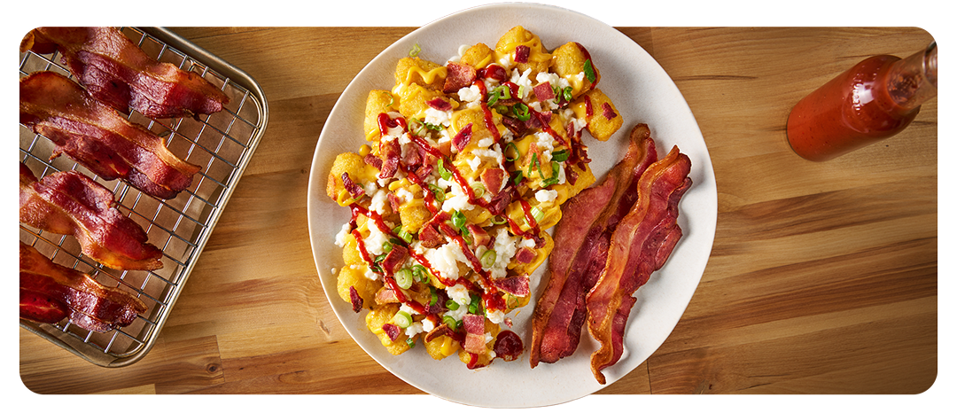A plate of potato tots mixed with chopped bacon, scrambled eggs and American cheese with a side of sliced bacon.