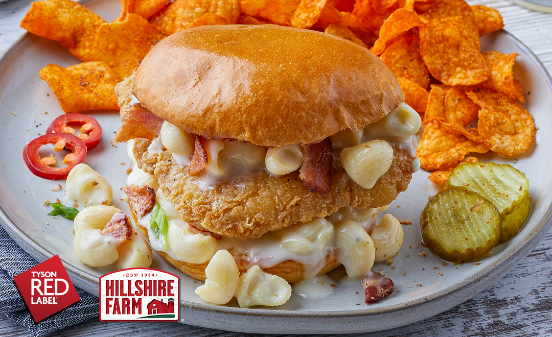 A plate with a breaded chicken breast covered in mac ‘n cheese and bacon crumbles in a soft bun   