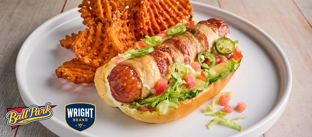 A white plate with a bacon-wrapped hot dog in a bun topped with tomatoes, avocado and sliced peppers with a side of waffle fries  