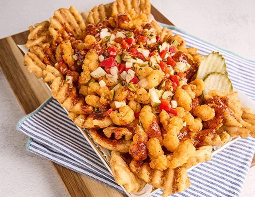 Waffle fries topped with crispy chicken bites, red sauce, relish, and pickles in a basket