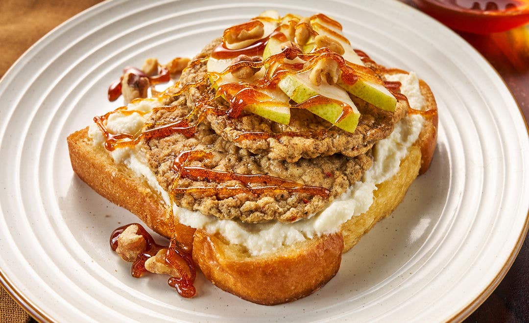A white plate with thick toast topped with two sausage patties, pear slices and walnuts, drizzled with honey