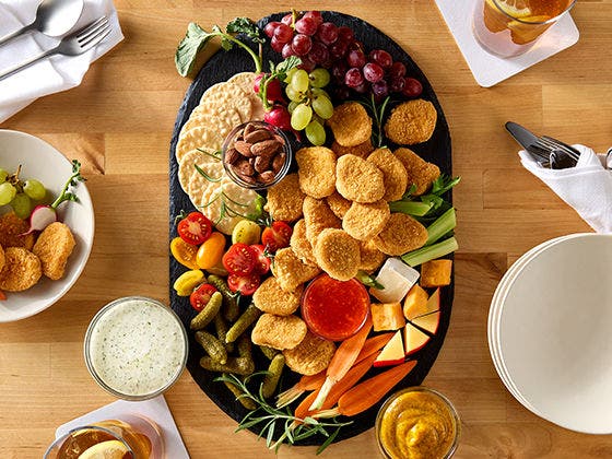Crispy gluten-free chicken nuggets on a board with fresh vegetables, fresh fruit, gluten free crackers and sauces on the side. 