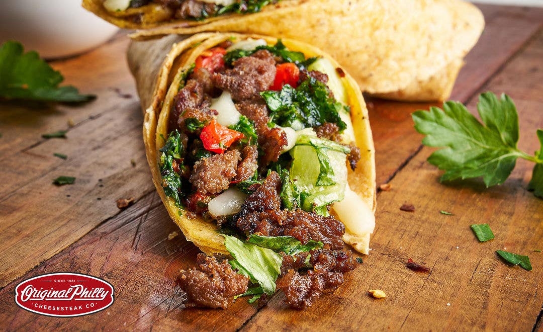 An open-ended steak wrap on a wood table with cooked sliced beef, roasted peppers and chimichurri in a flour tortilla   