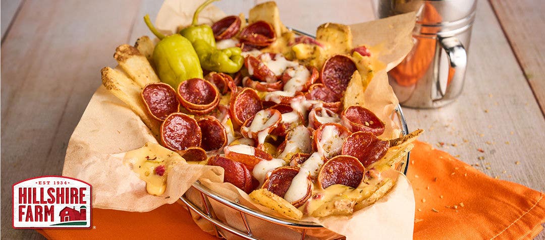 Fully loaded basket of French fries smothered in cheese and small, cupped pepperoni resting on a napkin on a wood table 