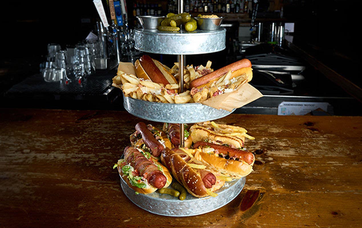 A three-tier serving tower on a wood tabletop with a variety of hot dogs, sides and sauces   