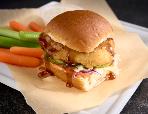 A plate with a small, breaded chicken filet in orange sauce with side of carrots and celery with dinner roll as bun