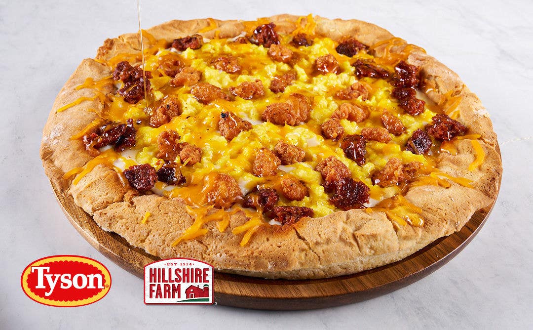 A wooden plate with a thick-crusted pizza loaded with scrambled eggs, sausage, chicken bites and cheese.  