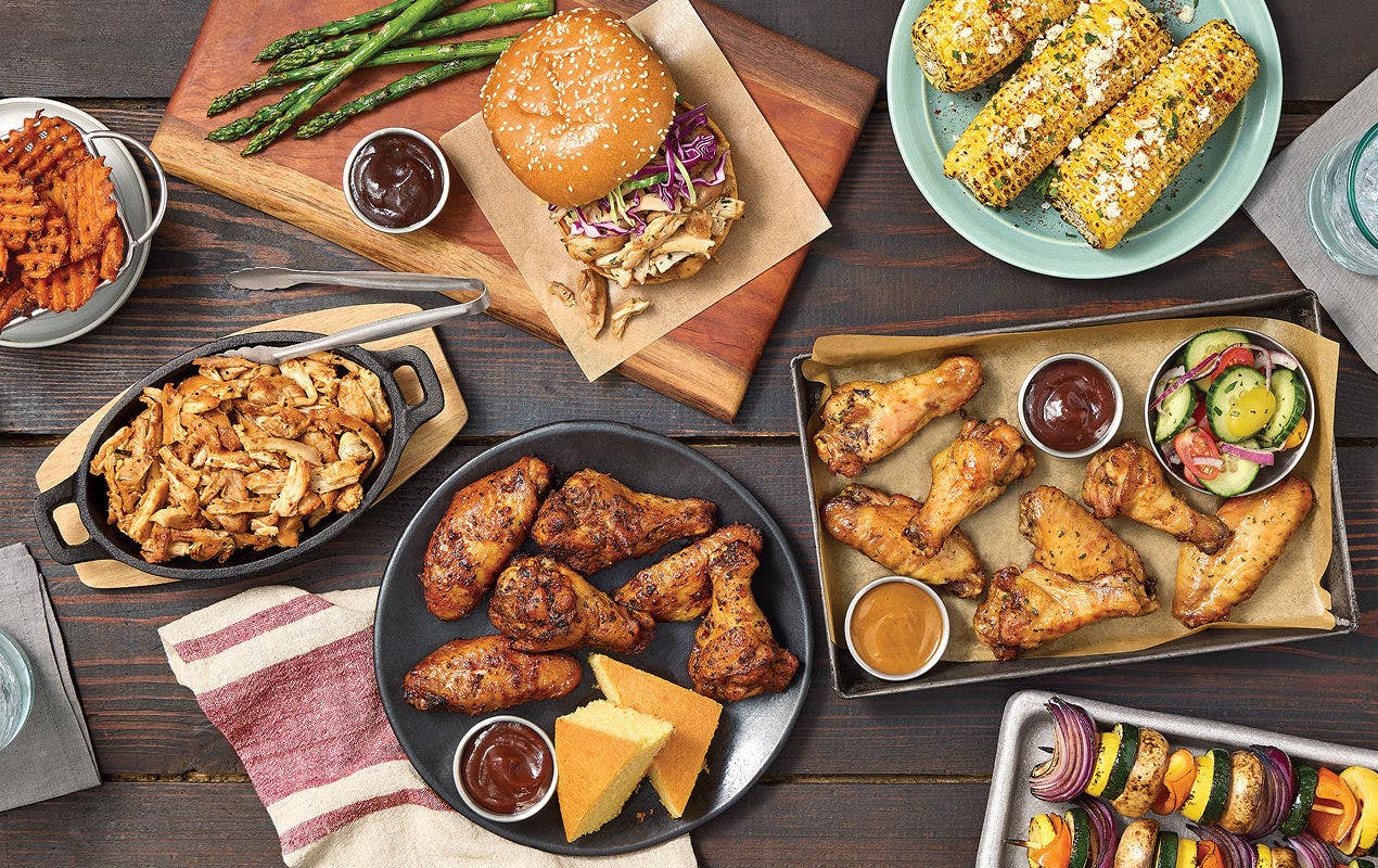 Overhead view of smoked chicken wings, a barbecue sandwich, grilled corn, asparagus, fries, sauces, and sides arranged on rustic wooden table