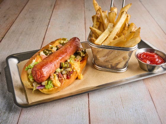 A loaded hot dog with cheese, onions, and relish on a metal tray with fries and ketchup