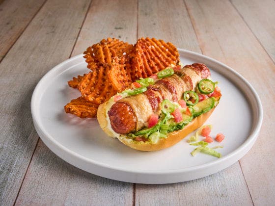 A bacon-wrapped hot dog topped with avocado, lettuce, tomatoes, and jalapeños, served with sweet potato fries