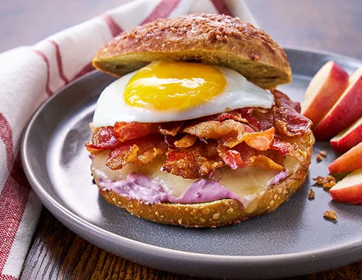 Crispy applewood smoked bacon breakfast bagel stacked with fried egg, white cheddar, and black currant cream cheese on a maple apple-cinnamon bagel