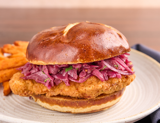 Crispy chicken schnitzel on a pretzel bun with mustard-braised red cabbage, sweet potato fries on a ceramic plate, set on a wooden table with a dark napkin