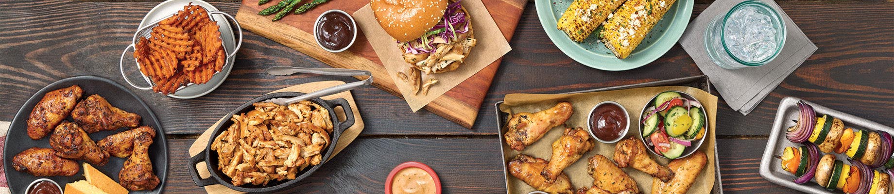 Overhead spread of Pitmaster smoked chicken dishes including wings, pulled chicken, sandwich, corn, fries, sauces, and grilled vegetables on wood table