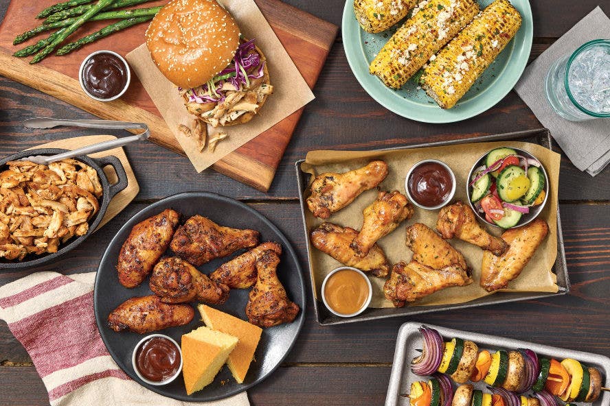 Two plates of Tyson® Pitmaster Chicken Wings and a sandwich featuring Tyson® Pitmaster Chicken Thigh Pieces, next to corn on the cob and kebabs