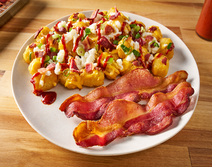 A plate with potato tots covered in cheese, bacon pieces, scrambled eggs, scallions and hot sauce with a side of bacon slices 