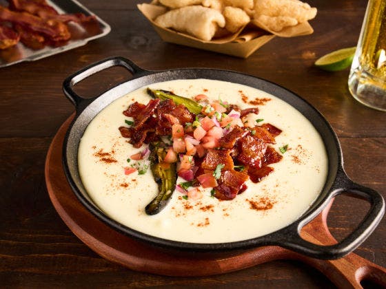 Skillet of creamy queso topped with bacon, pico de gallo, jalapeño, and chicharrones on the side