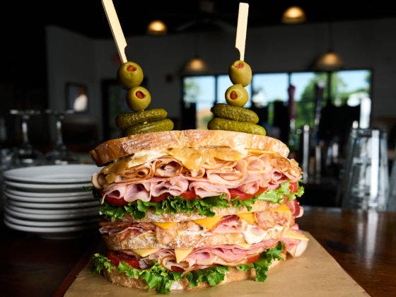 A tall, multi-layered sandwich with meats, cheeses, lettuce, tomato, with olives and pickles on skewers