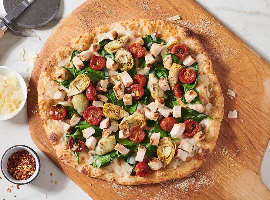 pizza peel holding a white sauce pizza with chicken, cheese, artichoke hearts and cherry tomatoes  