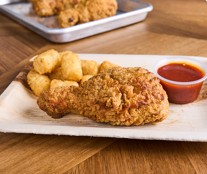 A breaded drumstick, potato tots and dipping sauce on a paper tray 