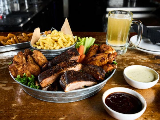 A large metal platter of barbecue with ribs, chicken wings, fries, celery, and carrots