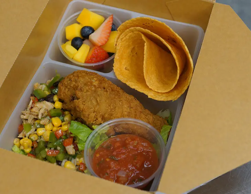 To-go container of breaded chicken drumstick served on rice, shredded lettuce, corn, black beans with fruit cup and tortilla 
