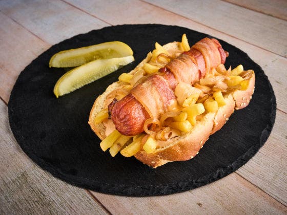 A bacon-wrapped hot dog topped with fries and grilled onions, served with pickles on a black plate